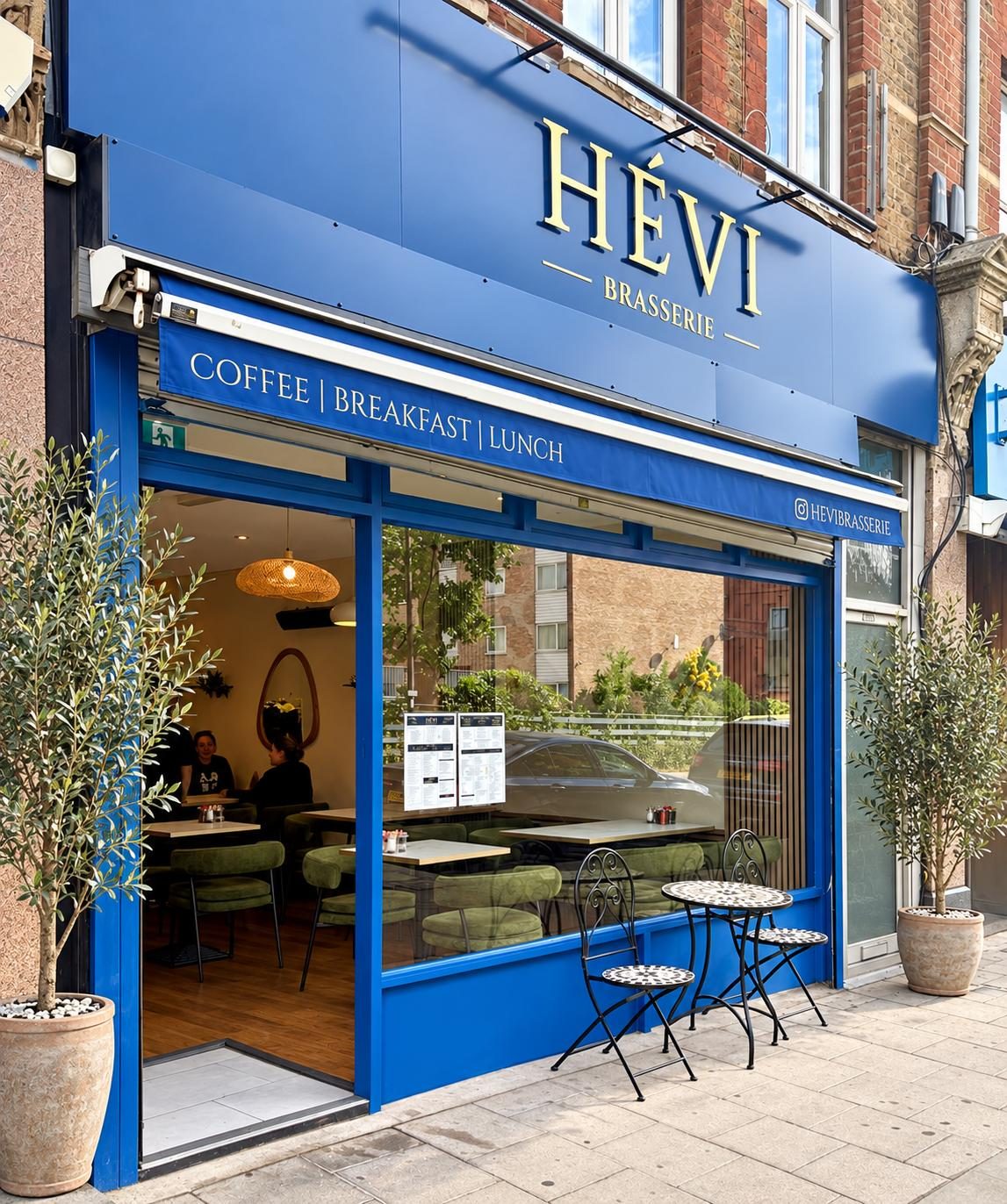 HÉVI Brasserie's blue shopfront on Green Lanes, with olive trees and a bistro table outside